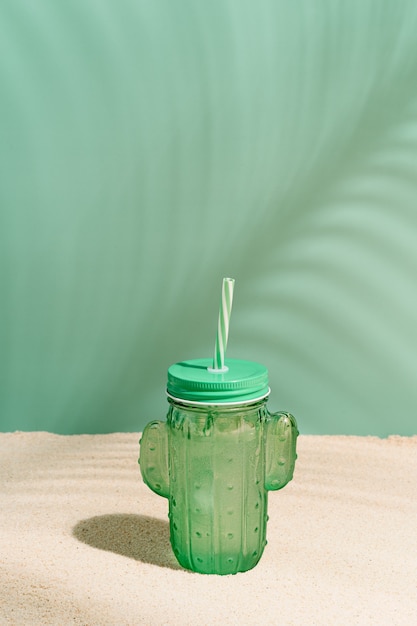 Copo refrescante com bebida em forma de cacto e palha na areia de uma praia tropical com sombras de folhas de coqueiro no verão