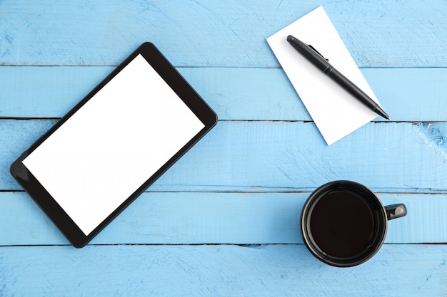 Copo, tablet, caderno de papel pequeno e uma caneta preta em uma madeira azul