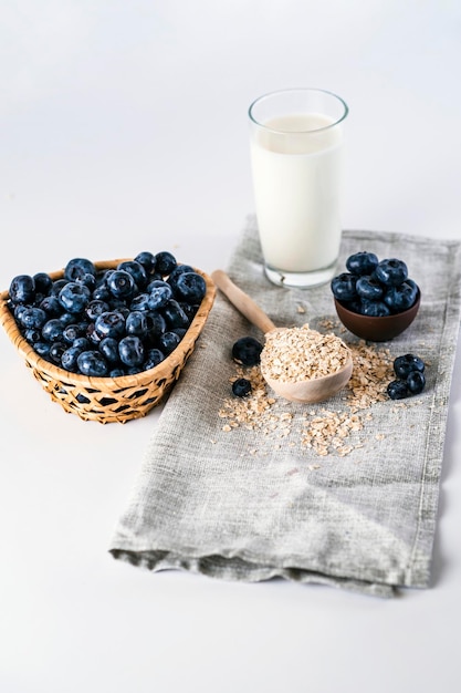 Copos de avena cruda con arándanos negros y un vaso de leche Alto ángulo de arándanos frescos y una cuchara de madera con avena colocada en la mesa cerca de un vaso de leche y sobre fondo blanco en la sala de luz