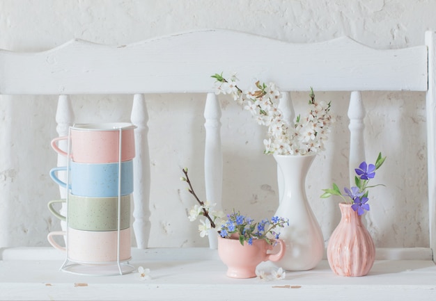 Copos e vasos com flores da primavera na prateleira branca de madeira vintage