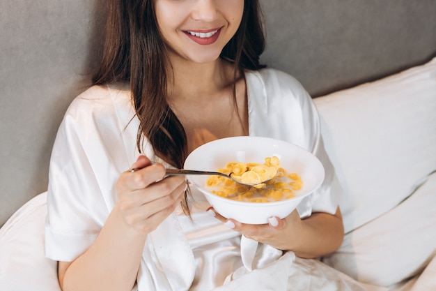 Copos de maíz para desayunar en la cama. hermosa mujer sosteniendo un plato de copos de maíz mientras está acostada en la cama.