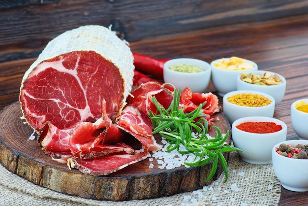 Coppa curada en rodajas con especias y una ramita de romero sobre fondo rústico de madera oscura.