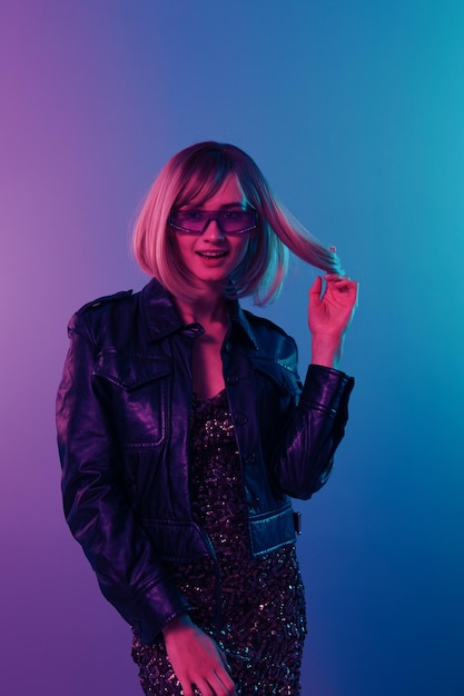 Coqueteando sexy sonriente hermosa mujer rubia en chaqueta de cuero vestido brillante gafas de sol de moda posando aislado en color azul rosa fondo de estudio de luz Fiesta de neón Concepto Cyberpunk Copiar espacio