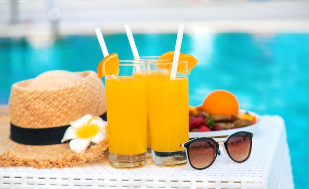 Coquetéis, óculos escuros, chapéu de palha e frutas frescas em uma tigela branca à beira da piscina