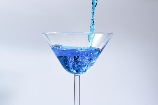 Coquetel com líquido azul em vidro Copo com água azul, derramando com líquido com salpicos e gotas Copo de Martini enchimento com álcool com salpicos no fundo branco Conceito de bebida refrescante