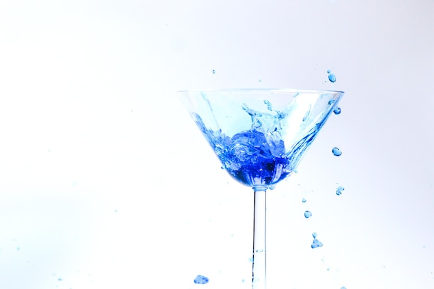 Coquetel com líquido azul em vidro. Copo com água azul derramando líquido com respingos e gotas. Copo de Martini enchendo-se de álcool com salpicos em fundo branco. Conceito de bebida refrescante.