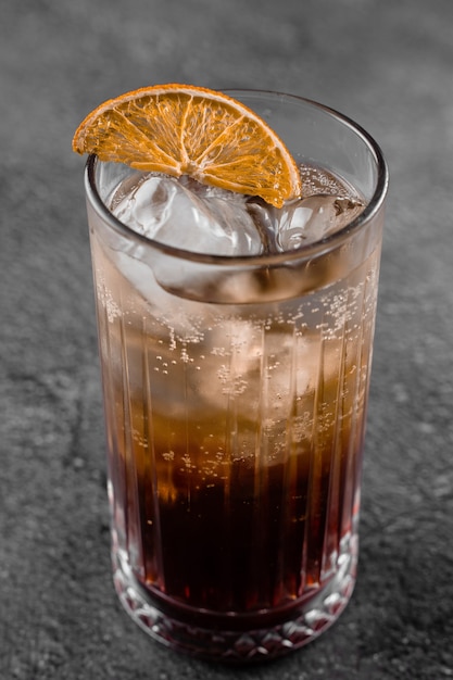Coquetel em copo com coca-cola, álcool, gelo, decorado com laranja seca na superfície cinza.