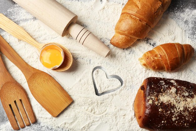 Coração de farinha, croissant e utensílios de cozinha de madeira em fundo cinza