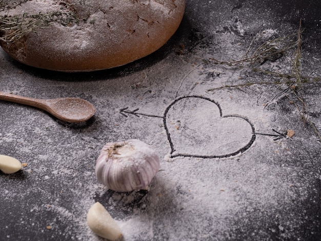 Coração desenhado na farinha sobre uma mesa cinza perto de especiarias e pão de alho caseiro.