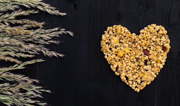 Coração disposto de granola e espigas em um fundo preto de madeira. postura plana. copie o espaço.