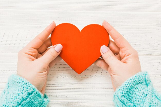 Coração vermelho nas mãos de uma garota em um suéter fechado em um fundo branco de madeira