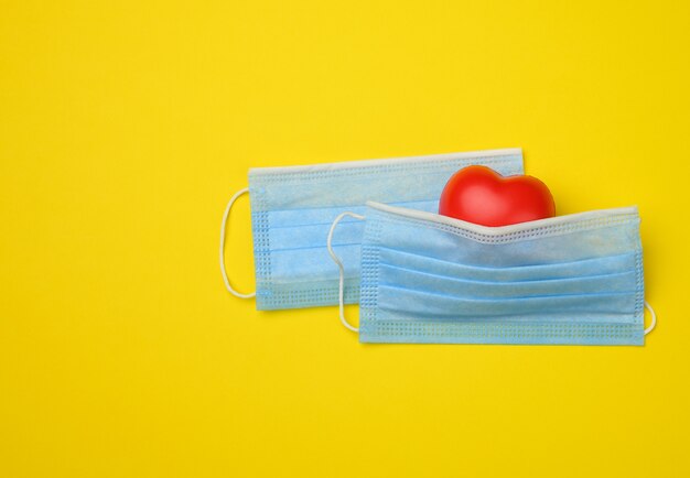 Coração vermelho repousa sobre uma máscara médica descartável branca, fundo amarelo, espaço de cópia