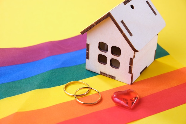 Corazón, anillos de boda y una pequeña casa de madera en la bandera lgbt, fondo amarillo, lugar de copia