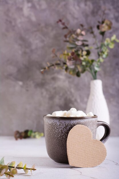 Corazón de cartón y una taza de cacao con malvaviscos en una taza sobre una mesa gris Vista vertical