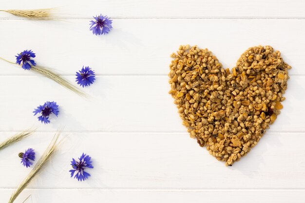 Corazón de granola, espiguillas y acianos sobre un fondo de madera blanca. Endecha plana. Copie el espacio.