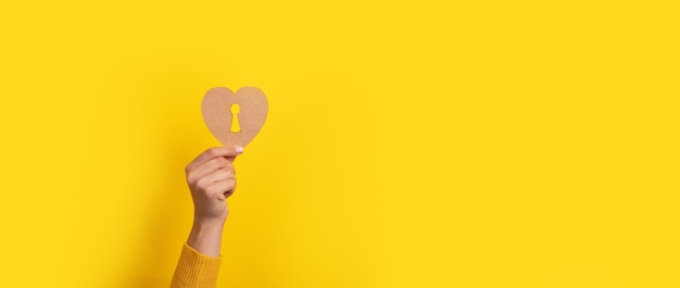 Corazón de madera con ojo de cerradura en la mano sobre fondo amarillo, imagen panorámica