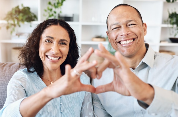 Corazón de la mano y retrato de una pareja madura con amor en el sofá para un vínculo romántico y feliz en la sala de estar Manos emoji y cara de un hombre mayor con una mujer feliz o sonriente mientras disfruta de la jubilación en casa