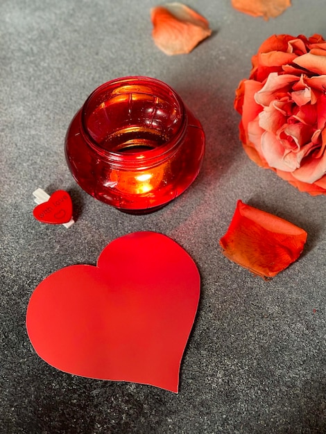 Un corazón de papel rojo brillante, una rosa roja con pétalos dispersos y una lámpara encendida sobre un fondo oscuro