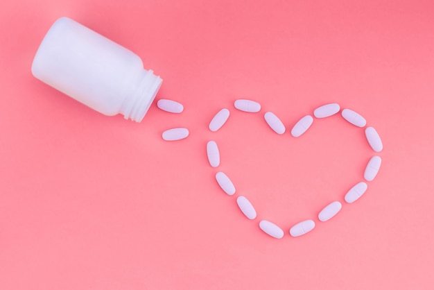 Corazón de pastillas sobre un fondo rosa. Las tabletas están esparcidas en forma de corazón de una lata blanca de medicina. Endecha plana.