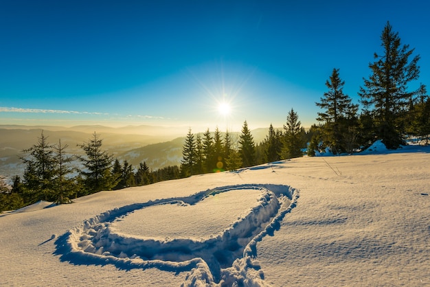 Corazón pisoteado en la nieve con los pies en el ventisquero en la ladera con una hermosa vista del bosque de coníferas