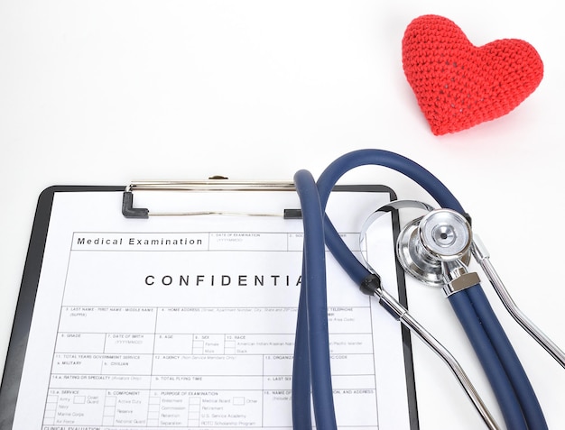 Foto corazón de punto rojo junto al estetoscopio y el formulario de examen del paciente aislado sobre un fondo blanco