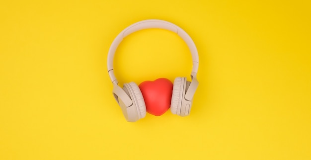 Corazón rojo y auriculares inalámbricos beige sobre fondo amarillo. Vista superior de gadget moderno