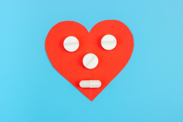 Corazón rojo con una cara hecha de pastillas sobre un fondo azul. El concepto de medicamentos, suplementos dietéticos, vitaminas para el tratamiento del corazón y la prevención de enfermedades.