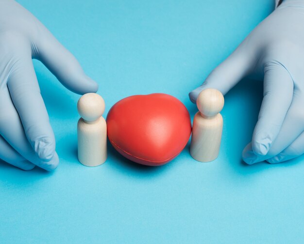 Corazón rojo y figurillas de madera de una familia, manos del médico en guantes azules, cerrar