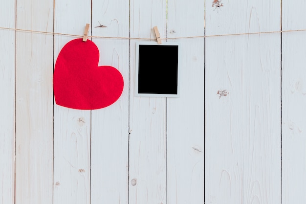 Corazón rojo y marco de foto en blanco colgando en cuerda para tender la ropa sobre fondo blanco de madera con espacio. Día de San Valentín.