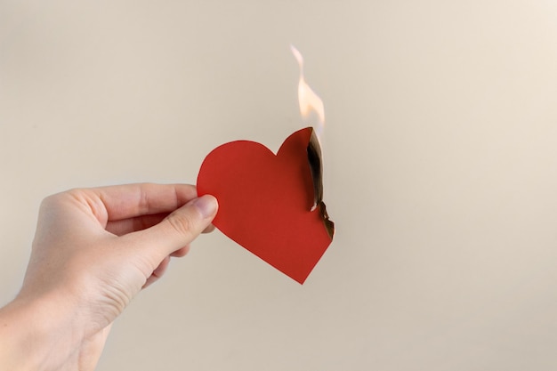 Un corazón rojo de papel arde en la mano de una mujer sobre un fondo beige. Concepto de amor infeliz, separación, sufrimiento.