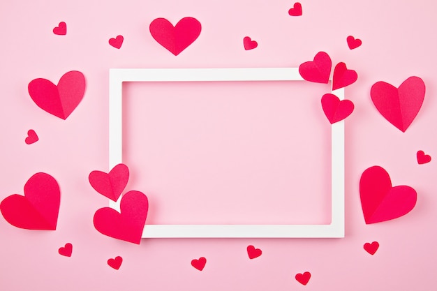 Foto corazones de papel y marco blanco sobre el fondo rosa pastel.