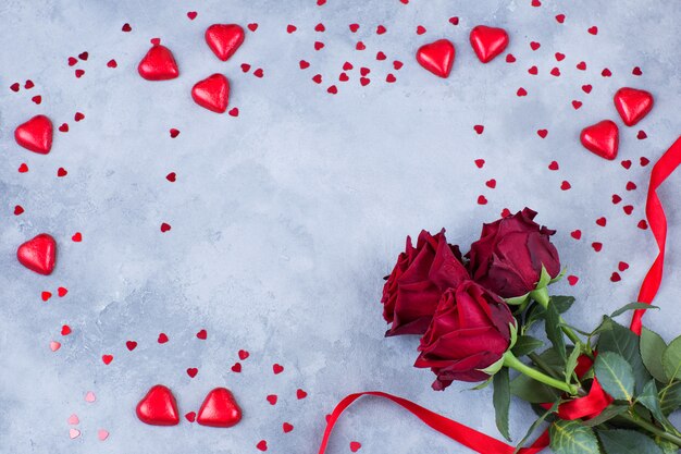 Corazones rojos de diferentes tamaños y tres rosas rojas atadas con una cinta sobre un fondo gris.