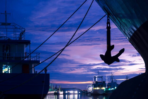 Corda de amarração de silhueta com âncora de navios de carga ancorados no porto contra o fundo do céu crepuscular