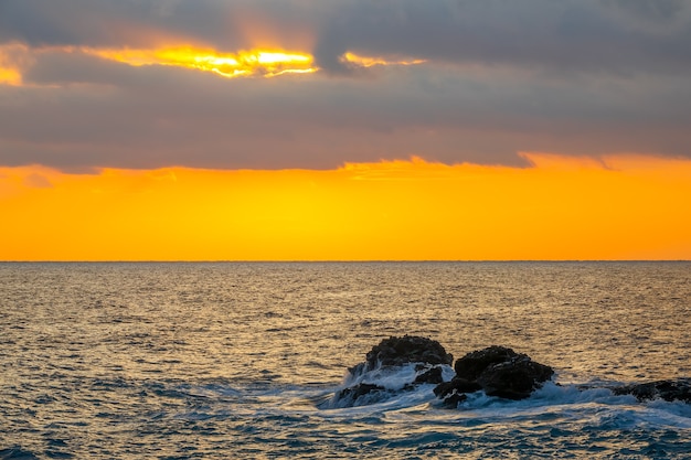 Cores do sol incríveis em céus nublados. Pedras costeiras no mar calmo