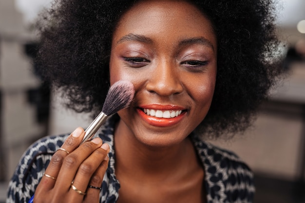 Cores lindas. mulher positiva de pele escura com cabelo encaracolado, fazendo um blush no rosto enquanto está sentada em frente ao espelho