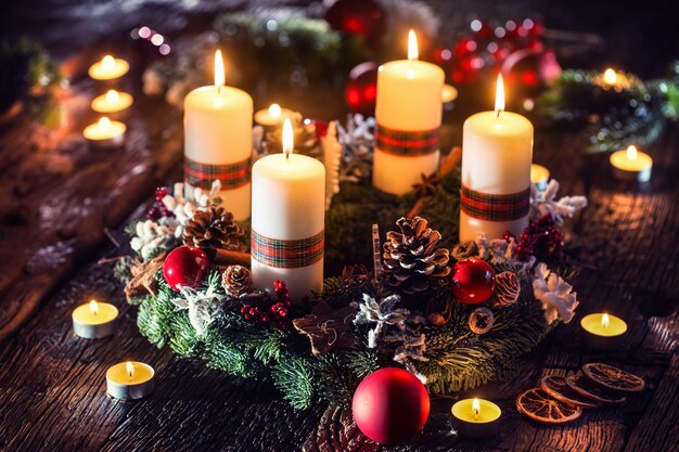 Corona de Adviento con cuatro velas blancas encendidas bolas de Navidad y adornos sobre un fondo de madera con ambiente festivo.