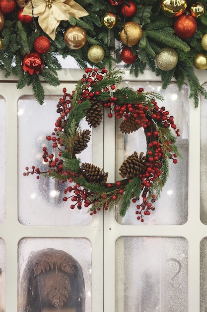 Corona navideña en la ventana con piñas y una guirnalda de adornos navideños y ramas.