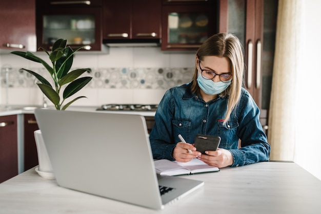 Coronavirus. Chica en la máscara de control de aplicaciones sociales en el teléfono. Mujer trabaja, aprende y usa la computadora portátil. Cuarentena. Quédate en casa. Persona de libre dedicación. Escritura, mecanografía. Concepto de comunicación y tecnología.