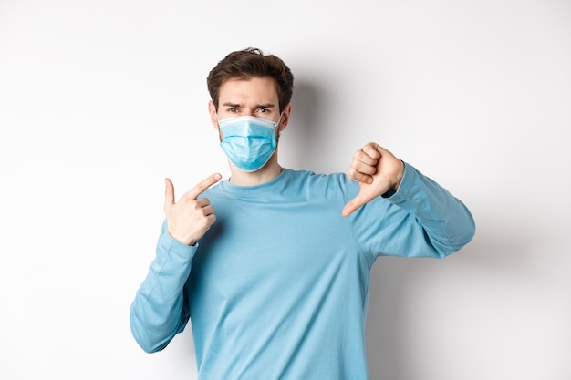 Coronavírus, conceito de saúde e quarentena. Homem zangado, apontando para sua máscara médica e mostrando os polegares para baixo, reclamando sobre fundo branco cobiçoso.