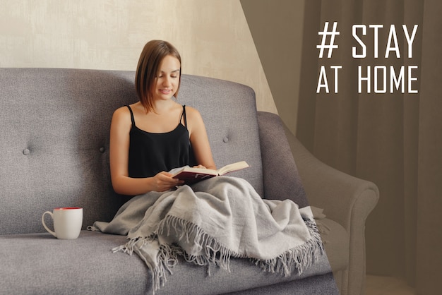 Coronavirus (COVID-19. Cuarentena, aislamiento. Quédese en casa durante la pandemia de coronavirus. Hermosa joven leyendo el libro, tumbado en el sofá envuelto en tela escocesa de lana. Relajarse en casa.