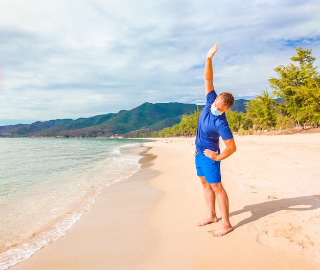 Coronavirus COVID 19 Joven hombre hermoso haciendo deporte realizando ejercicios de entrenamiento y gira cerca del mar por la mañana durante la cuarentena Vida activa en la protección quirúrgica de mascarilla facial esterilizante