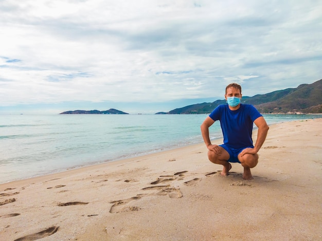 Coronavirus COVID 19 Junger schöner Mann, der Sport treibt, Trainingsübungen durchführt und morgens während der Quarantäne in der Nähe des Meeres hockt Aktives Leben im chirurgischen sterilisierenden Gesichtsmaskenschutz