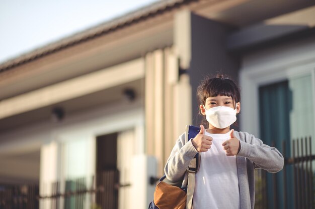 Coronavirus Covid-19 pm2.5.Online-Bildung.Kleines chinesisches Mädchen mit Gesichtsmaske zeigt Daumen hoch für Gutes und Glückliches zu Hause.