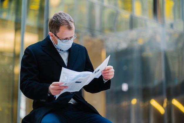 Coronavirus-Epidemie in der Stadt. Horizontale Aufnahme eines ernsthaften Mannes liest aufmerksam Zeitung, posiert vor verschwommenem Gebäudehintergrund, trägt eine Maske zur Verbreitung des Coronavirus. Geschäftsmann mit Presse