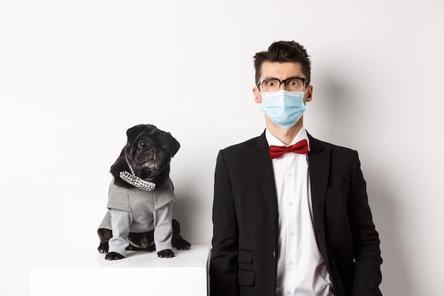 Coronavirus, Haustiere und Feierkonzept. Hübscher junger Mann und Hund, die Anzüge tragen, Kerl haben medizinische Maske, die über Weiß steht.