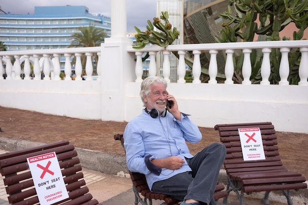 Coronavírus. Homem sênior sorridente relaxando ao ar livre sentado em um banco usando um telefone celular