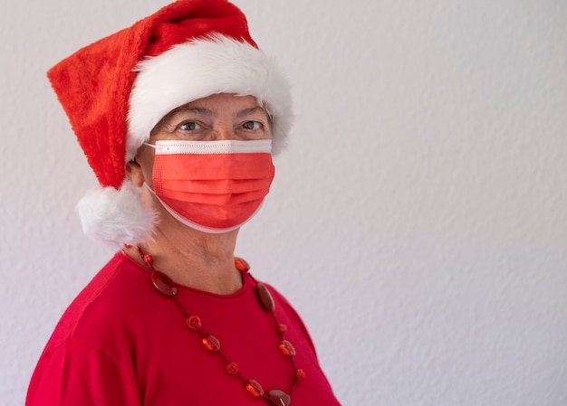 Coronavirus y Navidad 2020. Retrato de una mujer mayor con gorro de Papá Noel con mascarilla quirúrgica debido al coronavirus mirando a cámara. Color rojo sobre fondo blanco.