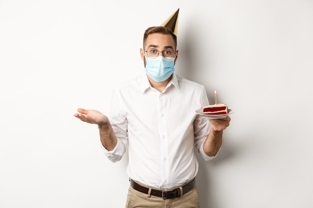 Coronavírus, quarentena e feriados. Homem confuso na máscara facial, segurando o bolo de aniversário e encolhendo os ombros, em pé sobre um fundo branco sem noção.