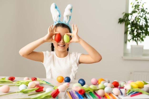 Coronavirus y Semana Santa. Niña linda con una máscara médica protectora con orejas de conejo con huevos de Pascua y flores de primavera.