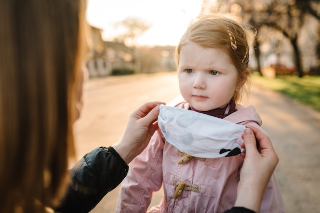 Coronavirus- und Luftverschmutzungskonzept. Kleines Mädchen und Mutter, die Masken tragen, gehen auf Straße. Mutter korrigiert Maskenkind. Symptome des Pandemievirus. Familie mit Kind im Freien. Krankheitsschutz.